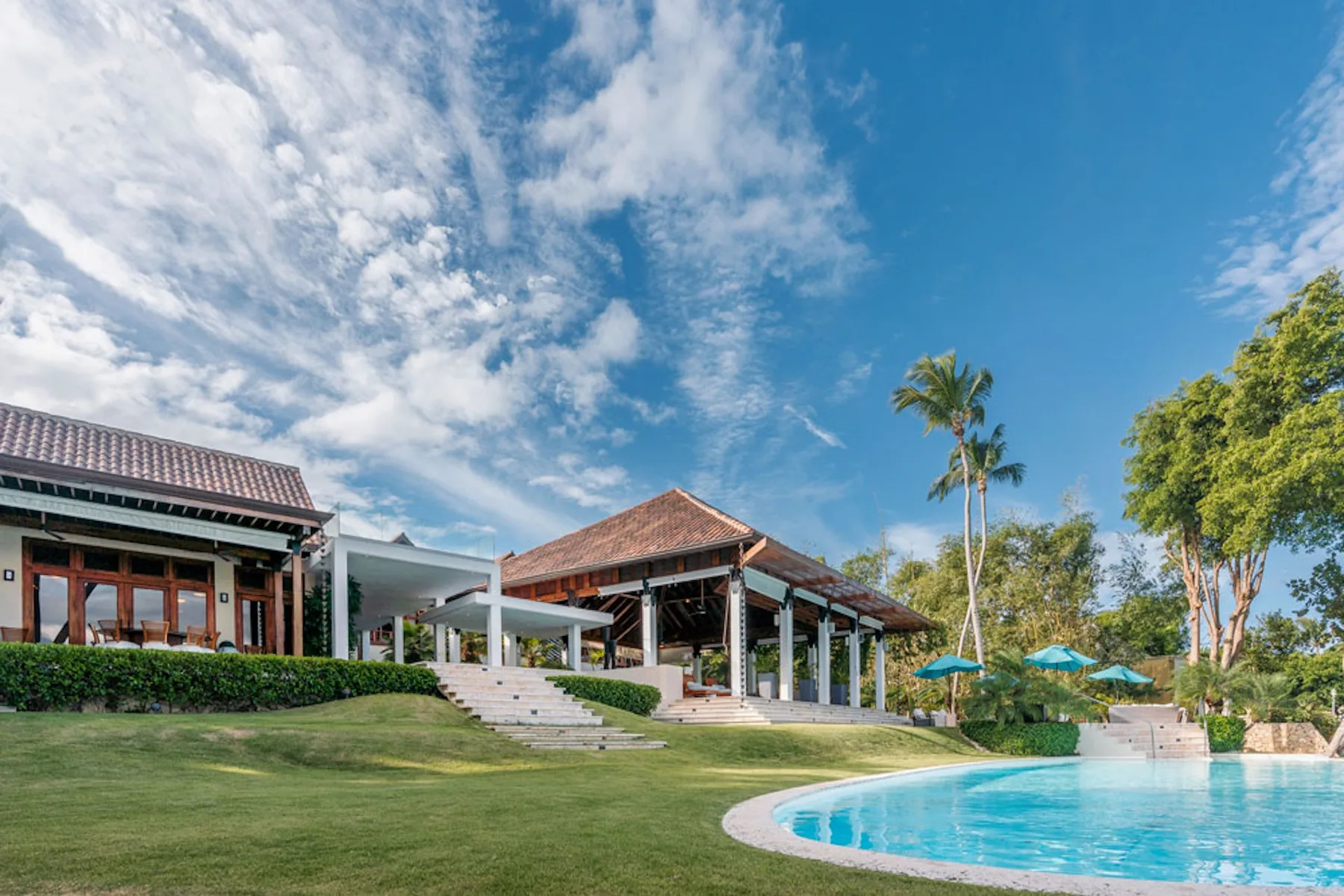 A landscaped outdoor pool sits beside a pavilion and low buildings under a blue sky.