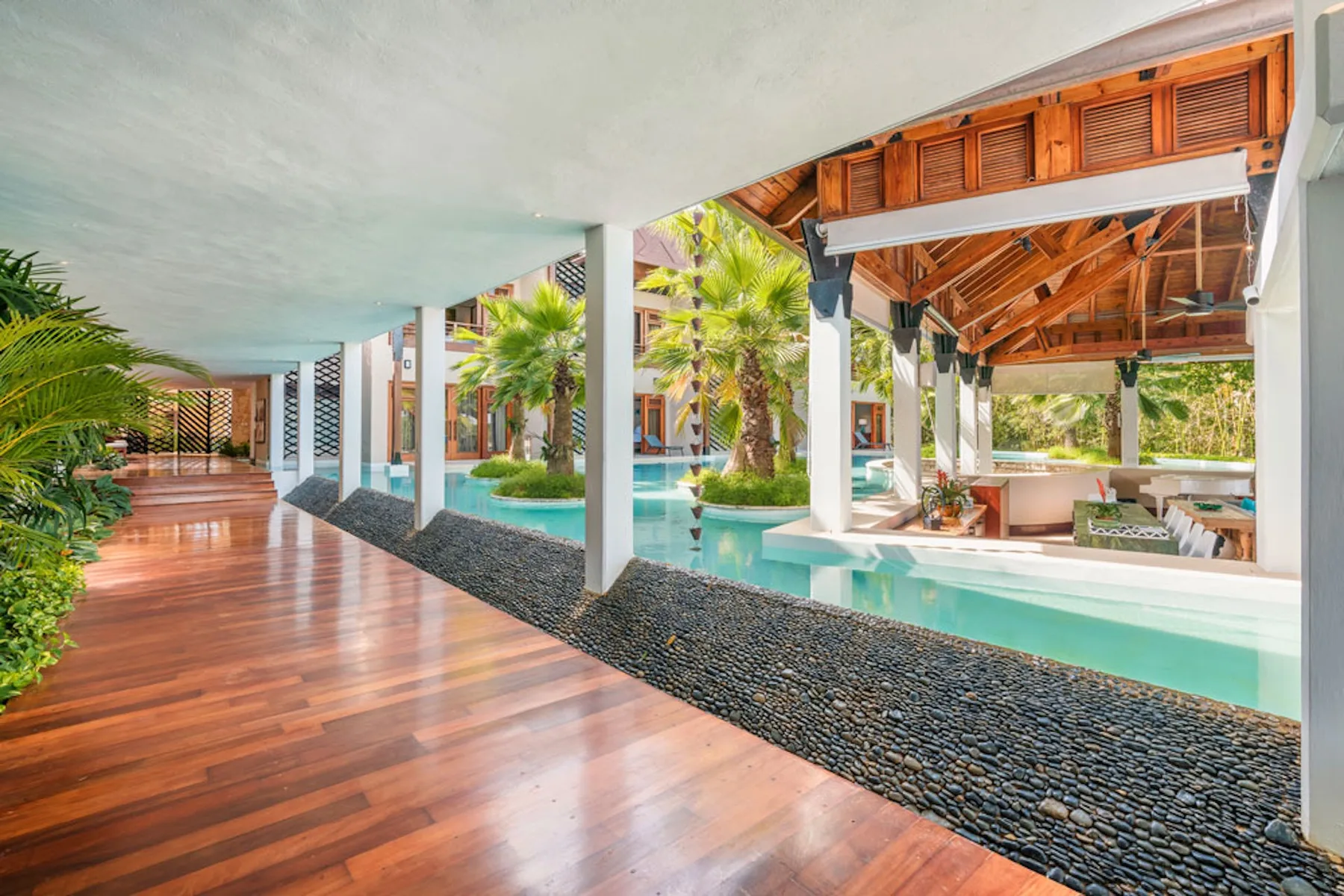 A covered wooden walkway runs beside a turquoise pool and tropical plants.