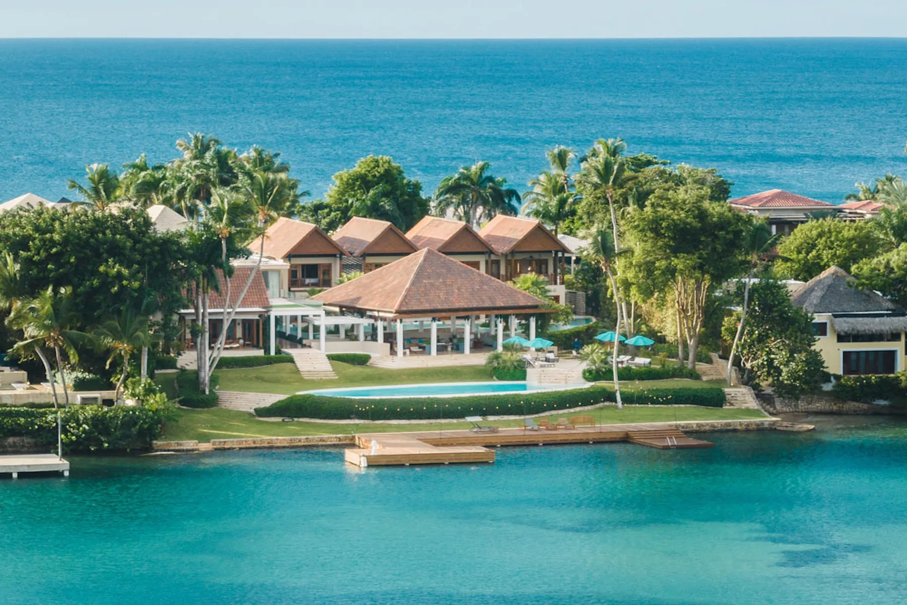 A tropical resort sits between turquoise water and the ocean, surrounded by palm trees.