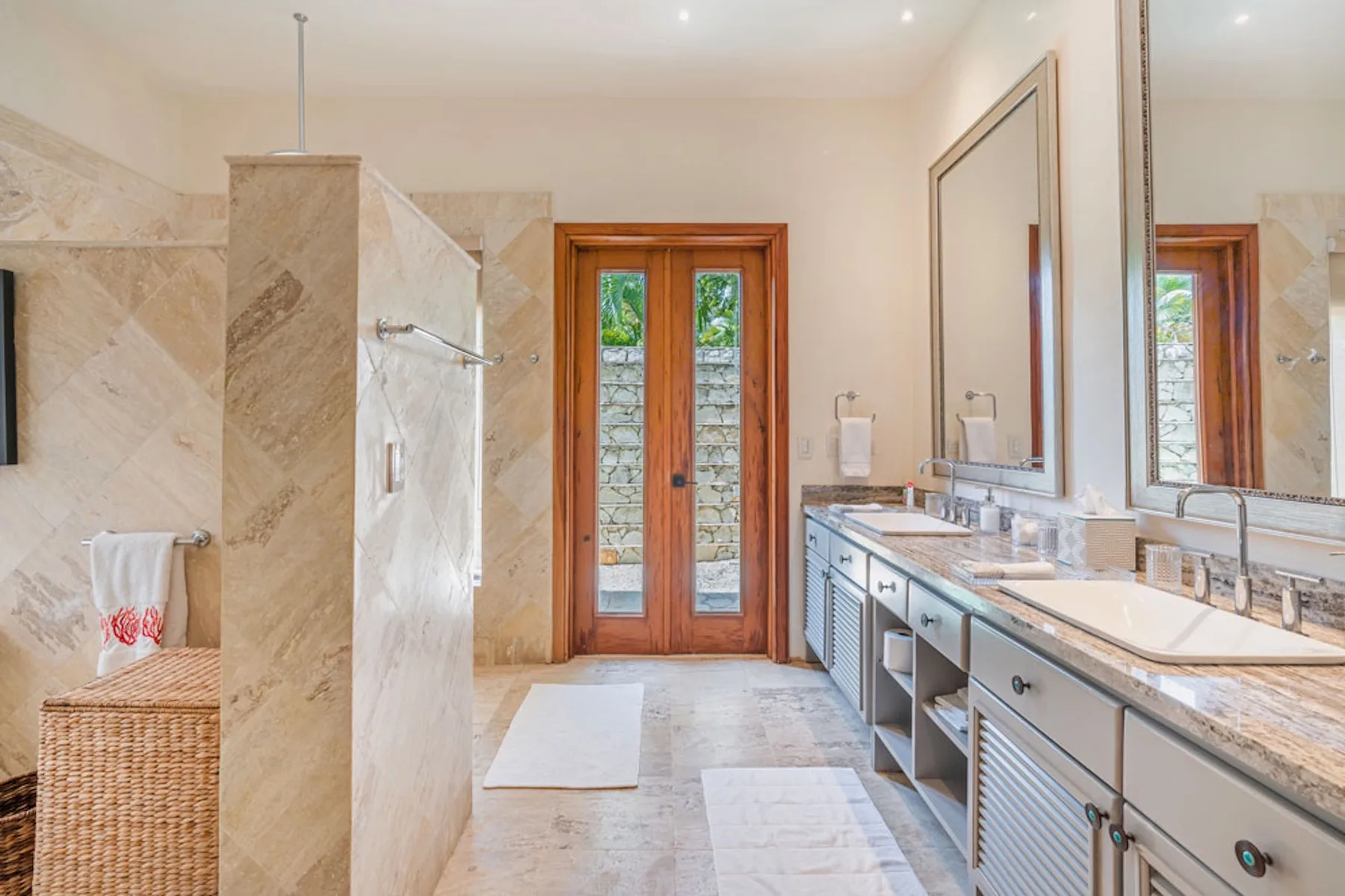 A bright bathroom with a glass shower, double vanity, and wooden doors.