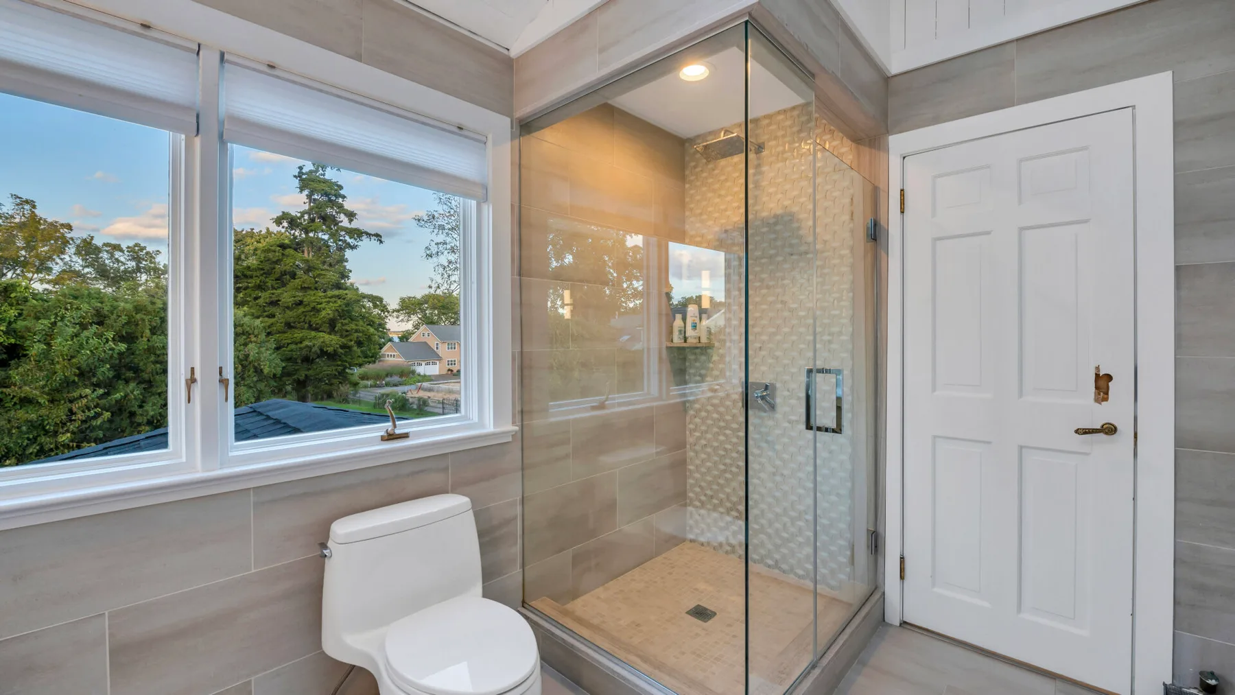 Un baño con ducha de vidrio, inodoro y una ventana con vista a los árboles.