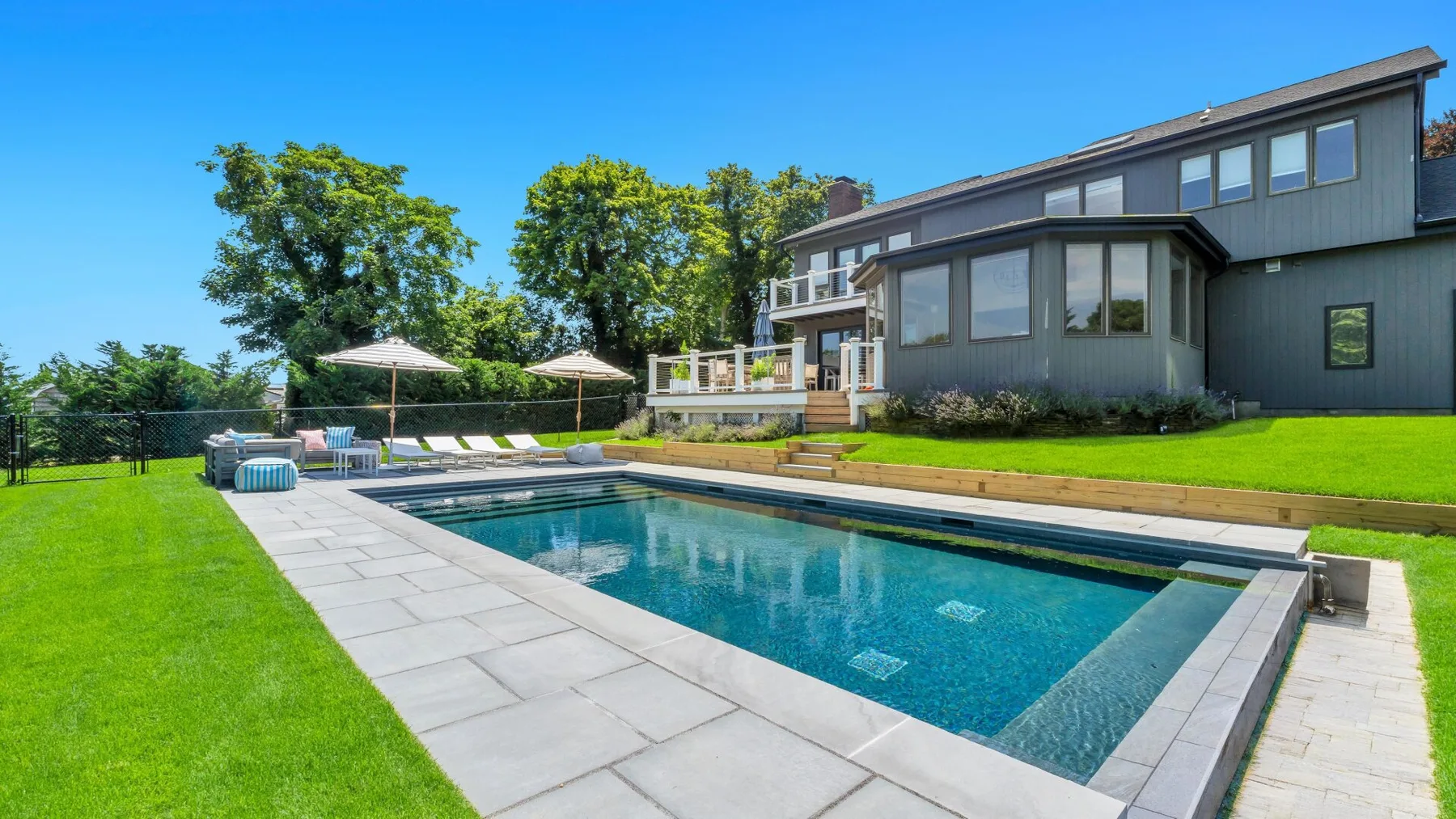 A backyard swimming pool is surrounded by a paved deck, lawn, and trees.