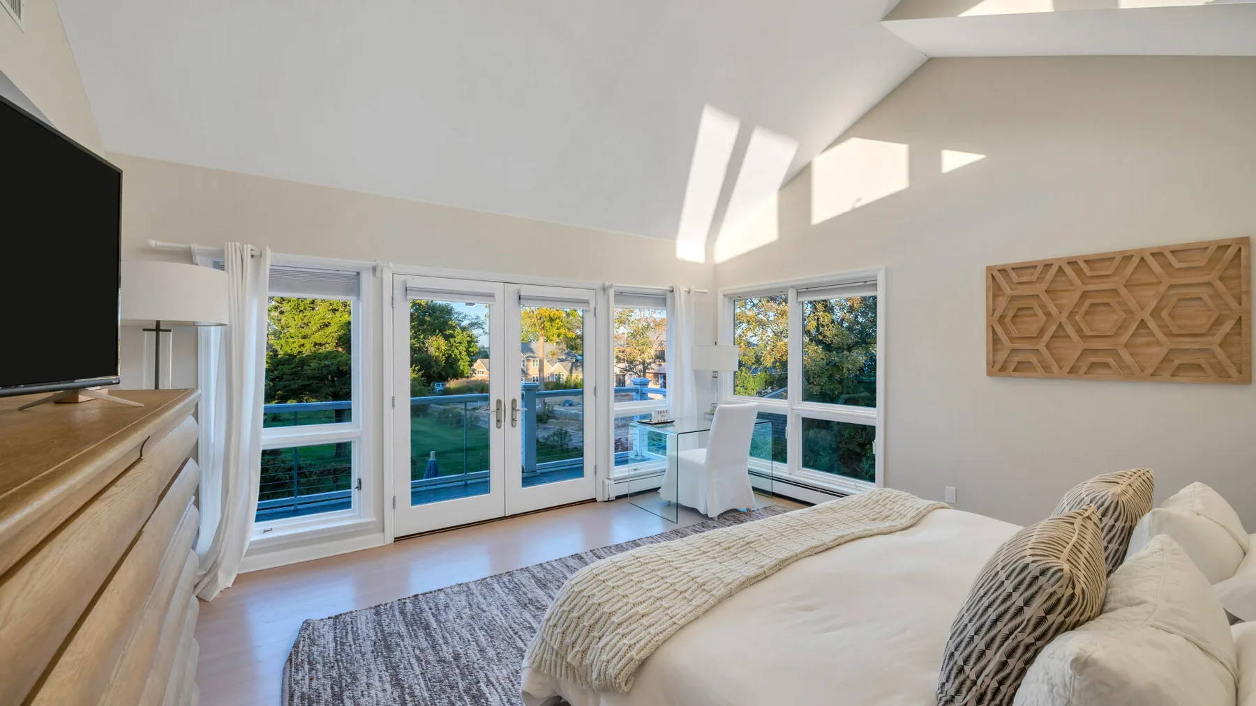 Un dormitorio luminoso tiene una cama junto a grandes ventanales con vistas a vegetación y una piscina.