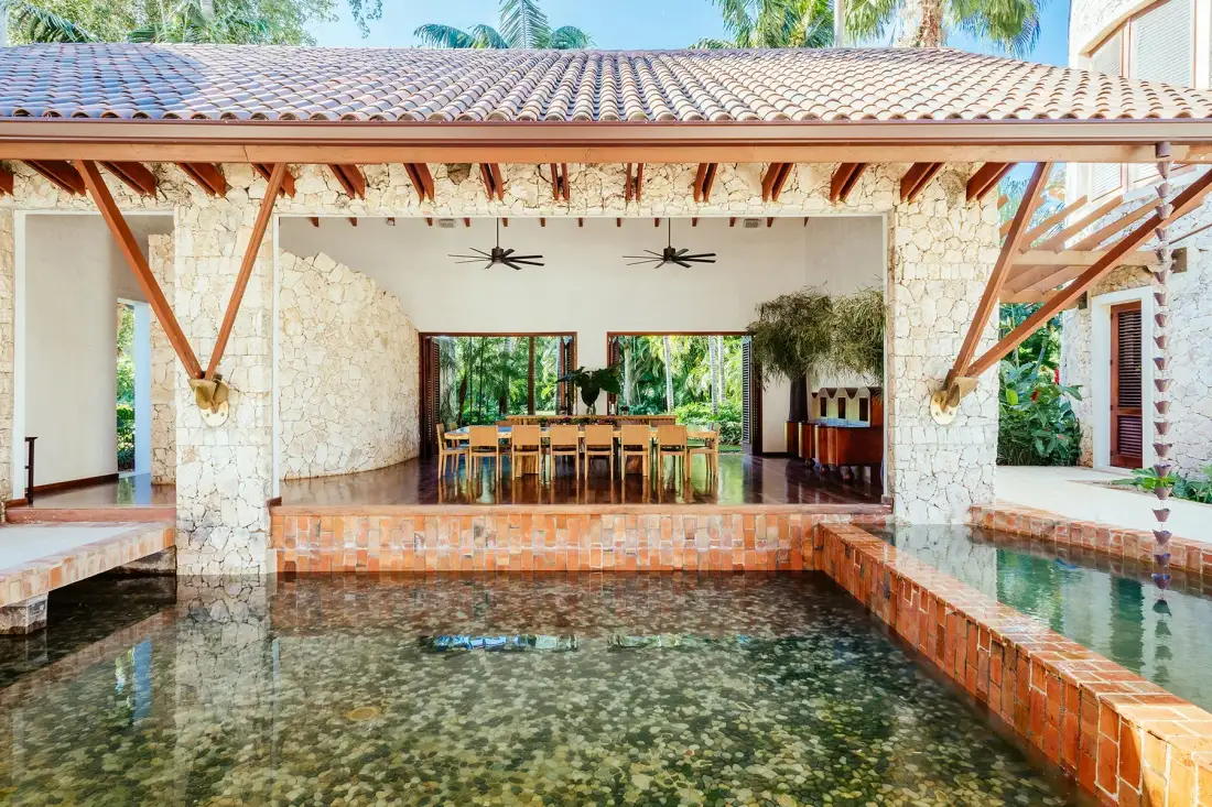 An open-sided dining area overlooks a swimming pool, with a long table beneath a tiled roof.