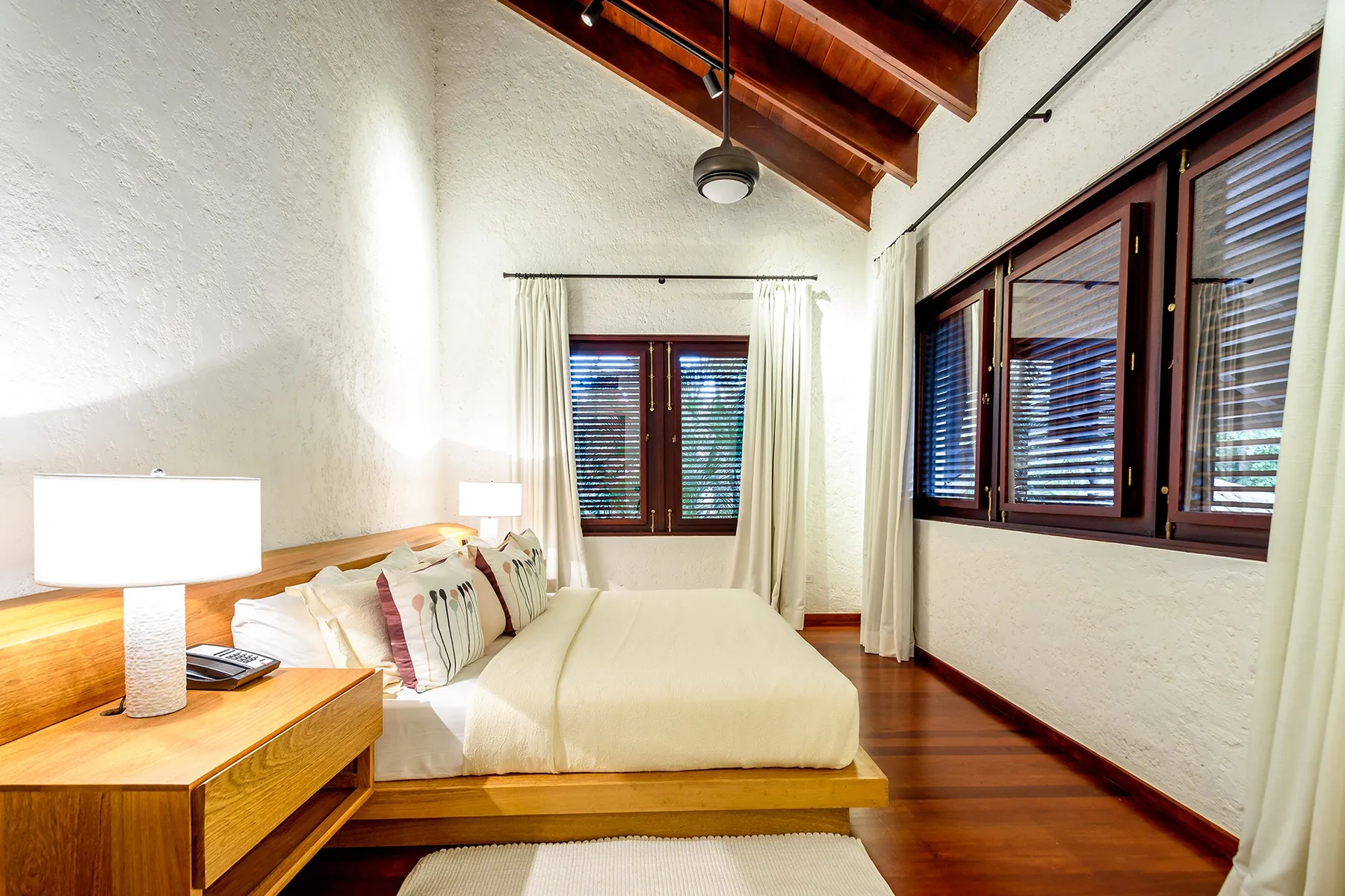 A bedroom with a bed beneath a sloped wooden ceiling and windows along the wall.