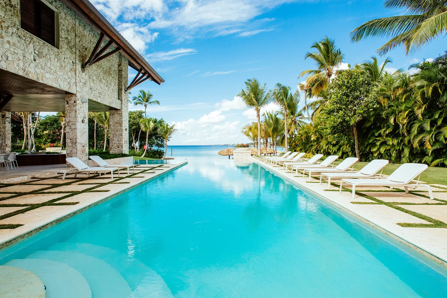 A long outdoor pool beside a stone building, with lounge seating and palm trees.