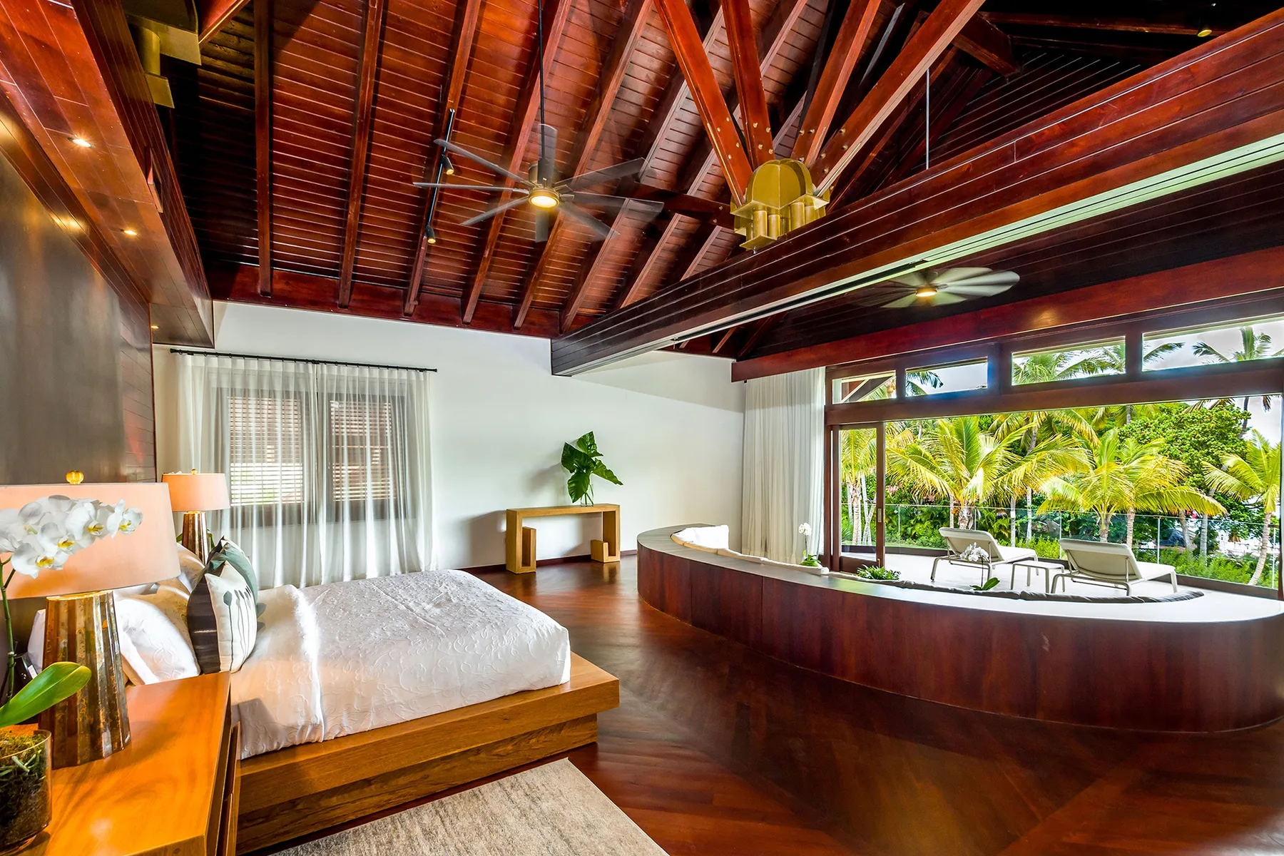A bedroom with a wooden bed beneath a high exposed timber ceiling, overlooking a tropical garden.