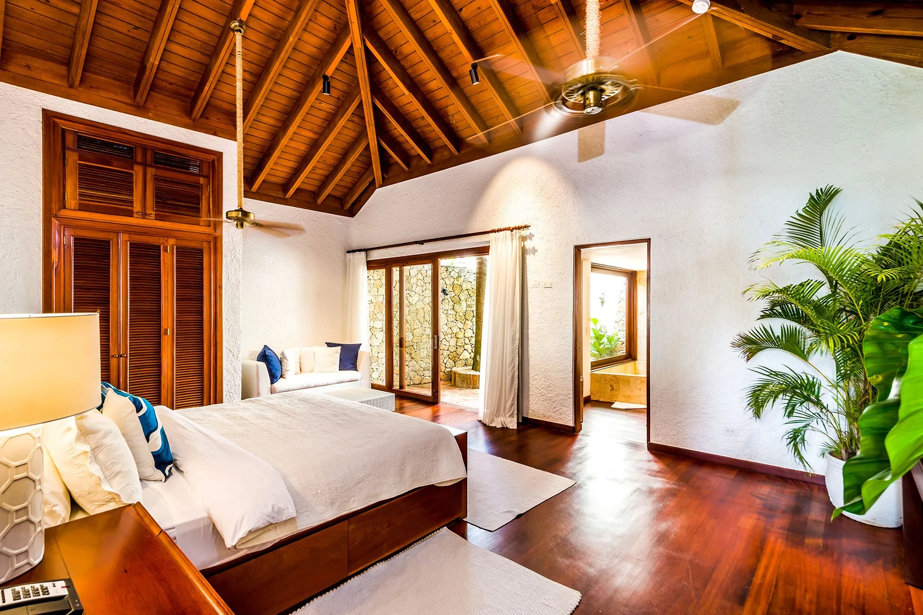 A bright bedroom with a large bed, wooden ceiling, and leafy plants.