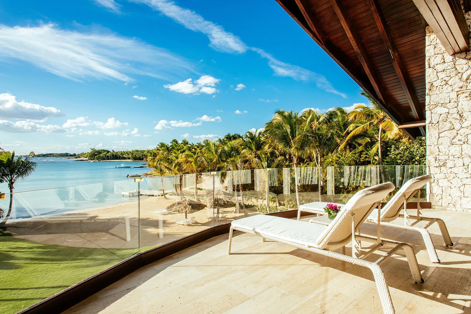 A sunlit terrace with lounge seating overlooks a tropical beach and calm water.