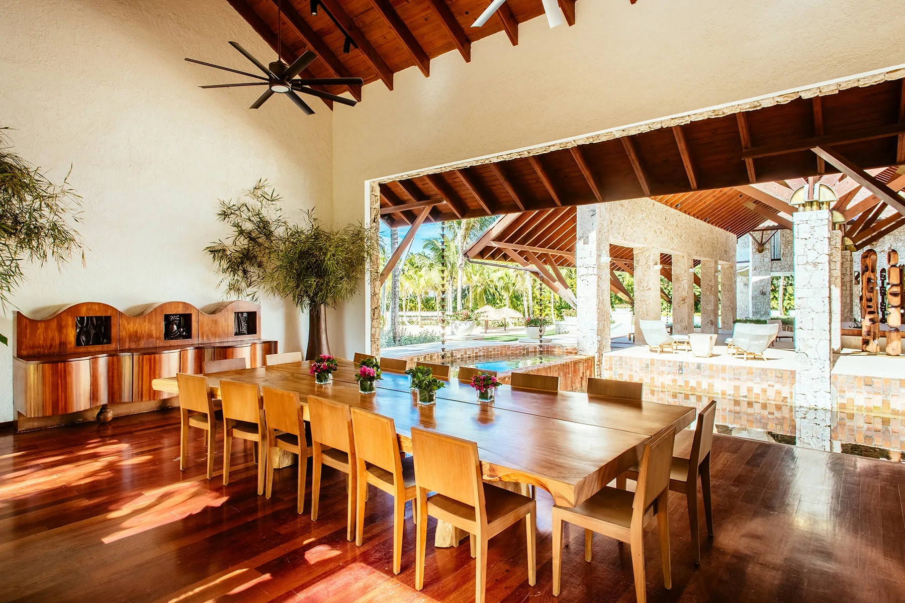 An open dining area with a long wooden table overlooking a pool and covered patio.