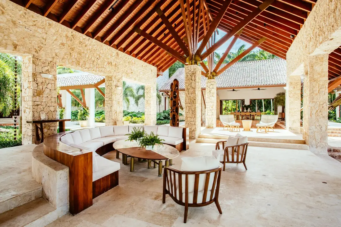 An open-air lounge has curved seating and wooden chairs beneath a high timber roof.