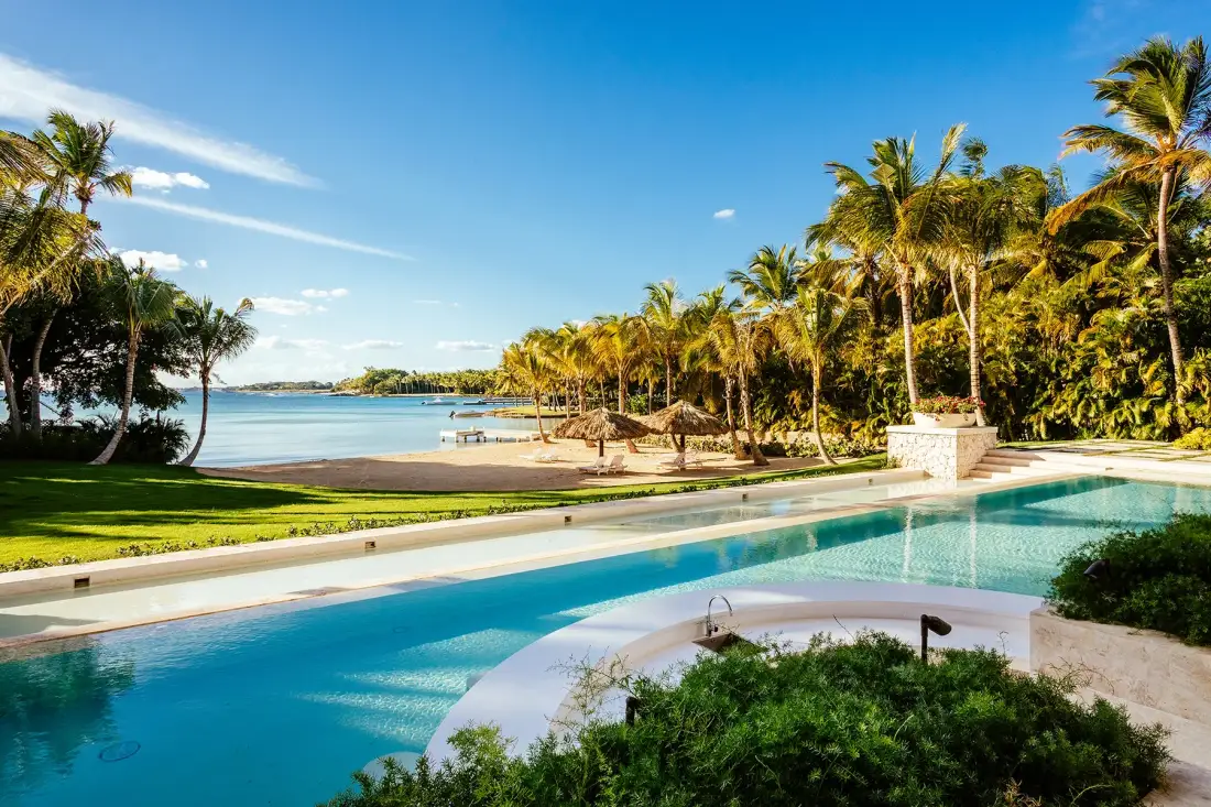 A swimming pool overlooks a sandy beach and palm-lined shoreline.