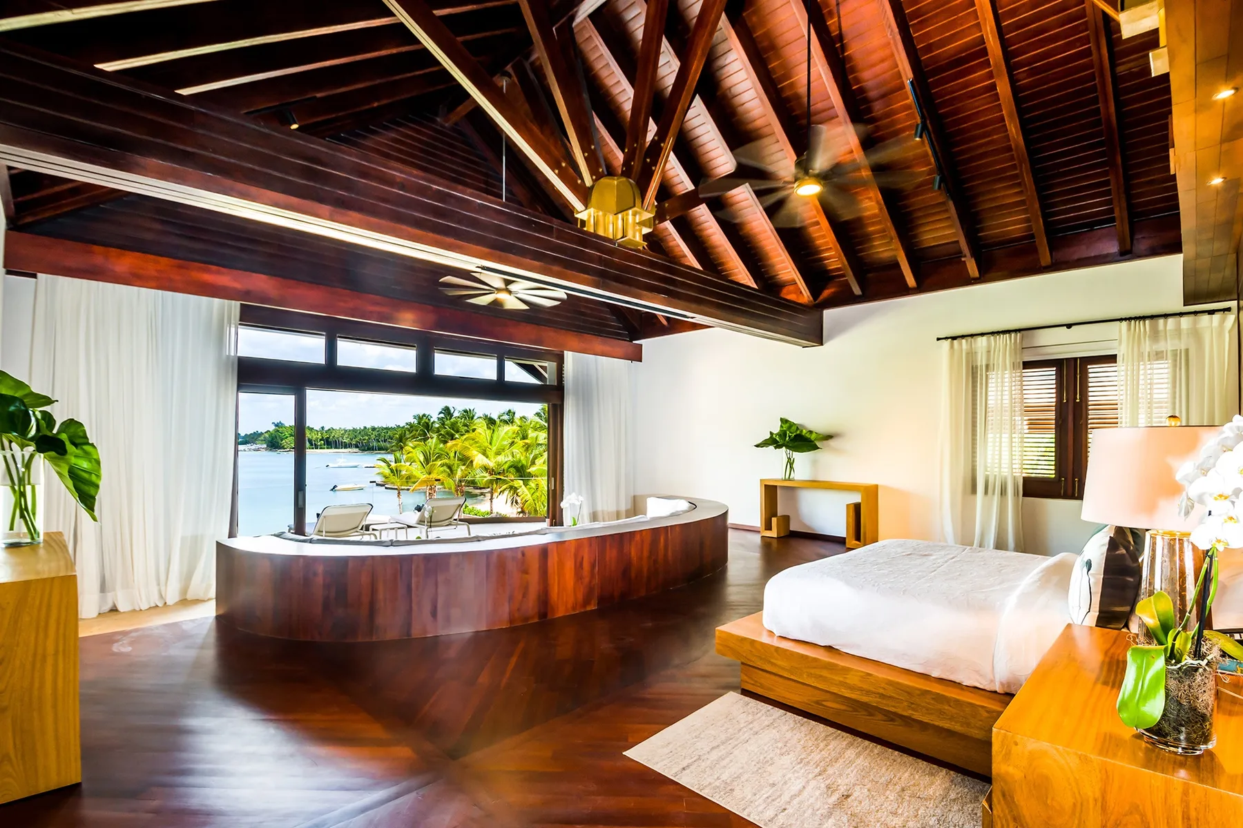 A bedroom with a curved wooden desk, white bedding, and a view of palm trees.