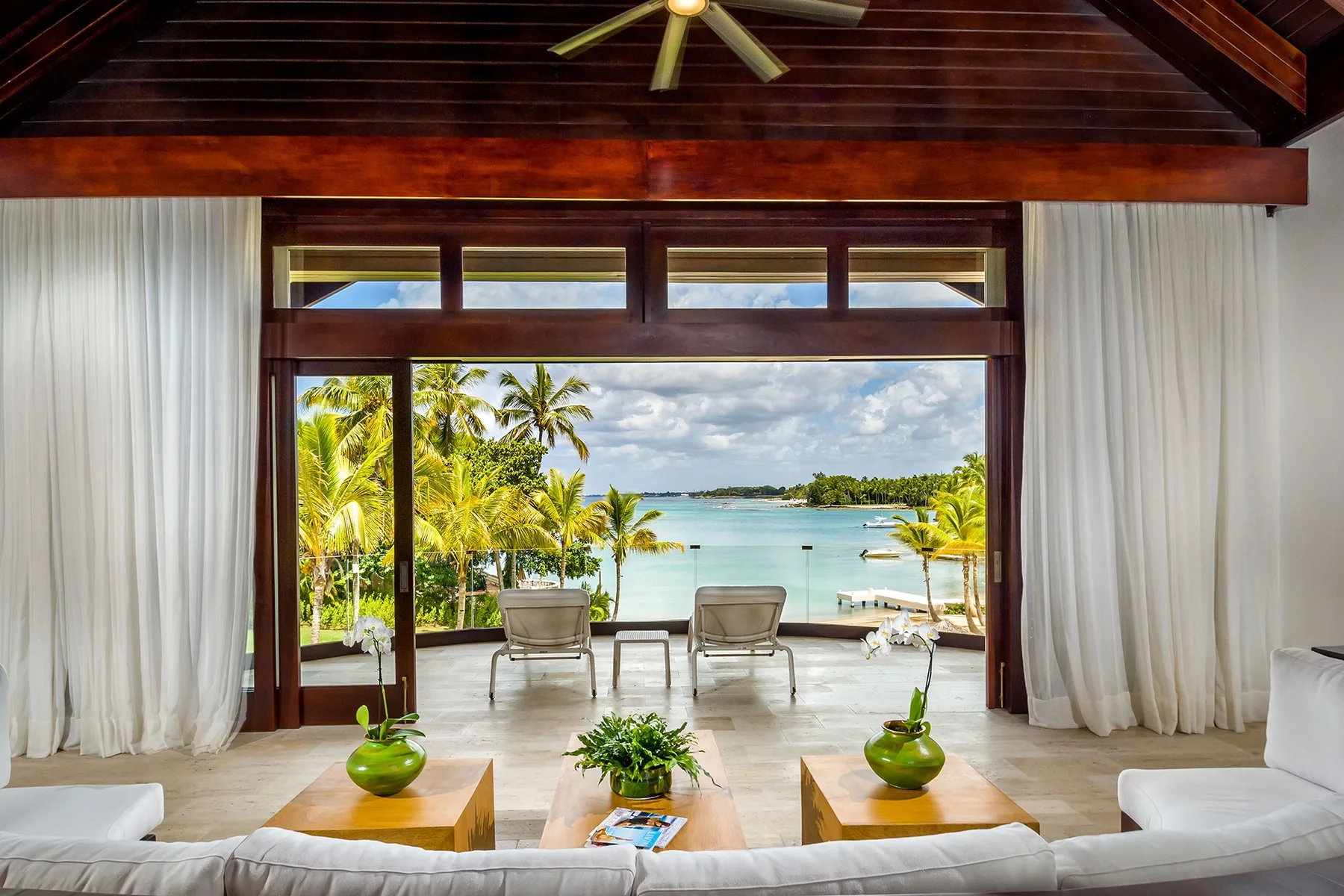A bright living area opens onto a tropical waterfront view, with seating beside the windows.