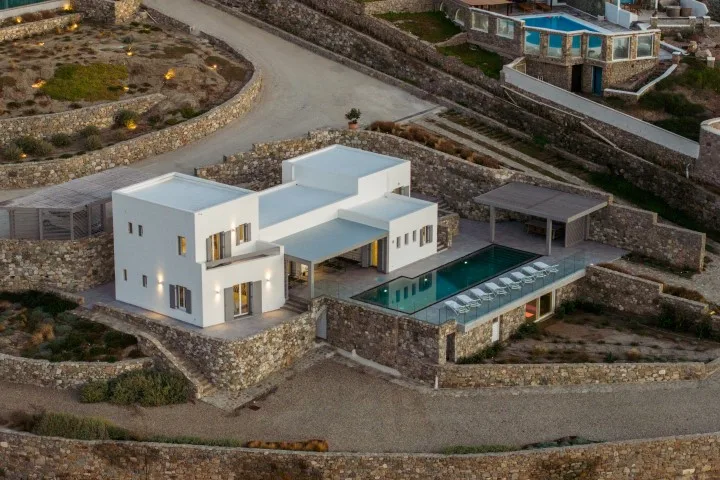 A white villa with a long swimming pool sits among stone walls.