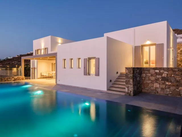 A modern white villa stands beside an illuminated swimming pool at dusk.