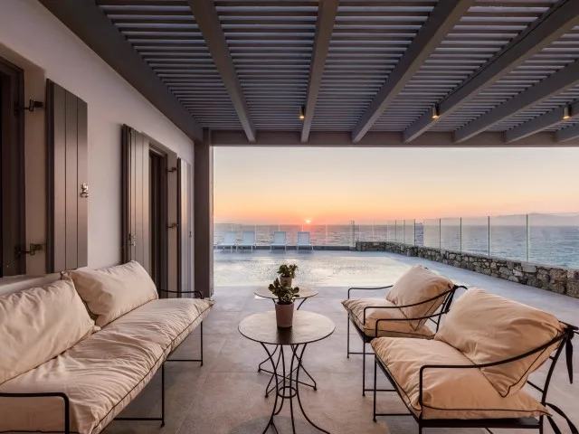 A covered terrace with cushioned seating and a view of the sea at sunset.