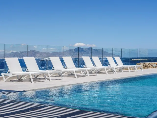 Una piscina junto a una fila de tumbonas blancas y una barandilla de vidrio.