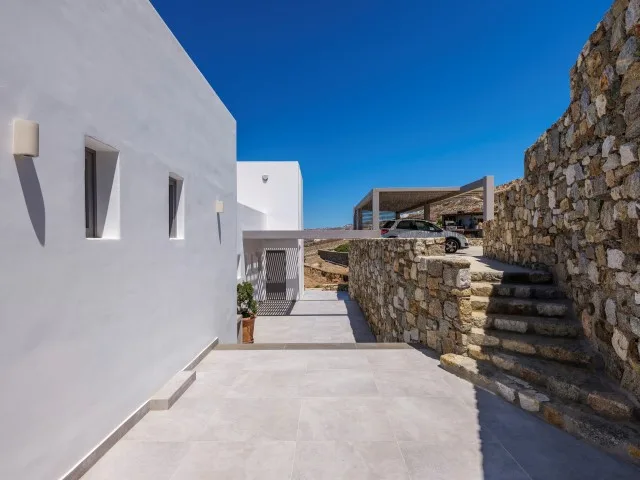 Un pasillo exterior pavimentado discurre entre un edificio blanco y muros de piedra.