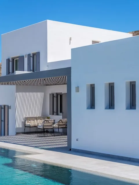 A modern white house beside a swimming pool, with a shaded outdoor seating area.