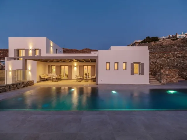 A modern house with a lit patio beside a swimming pool at dusk.
