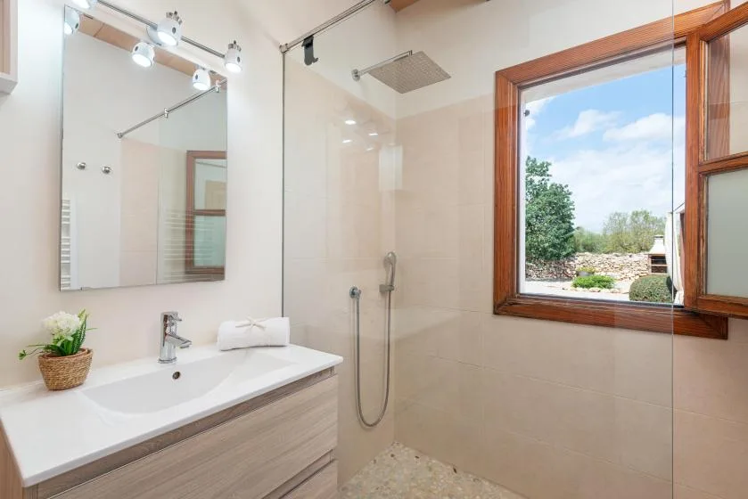 A bright bathroom with a sink, mirror, and glass-enclosed shower beside a window.