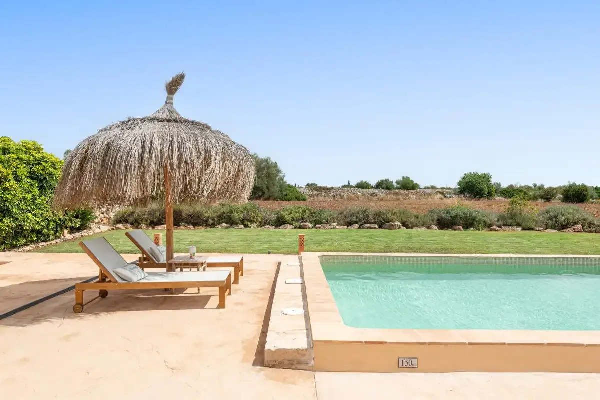 An outdoor swimming pool beside a paved patio with reclining seating and a thatched parasol.