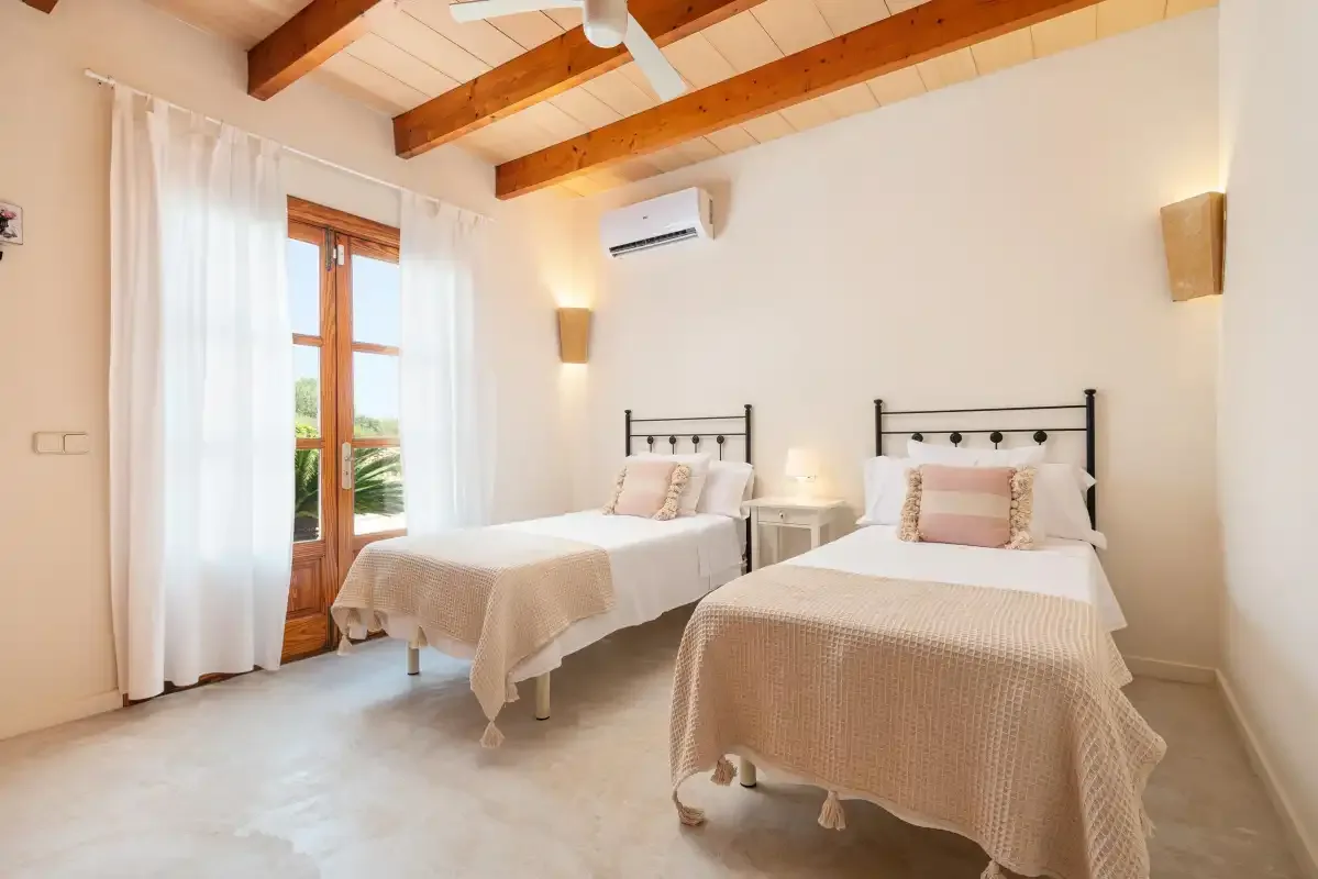 A bright bedroom with two beds and a tall window with white curtains.