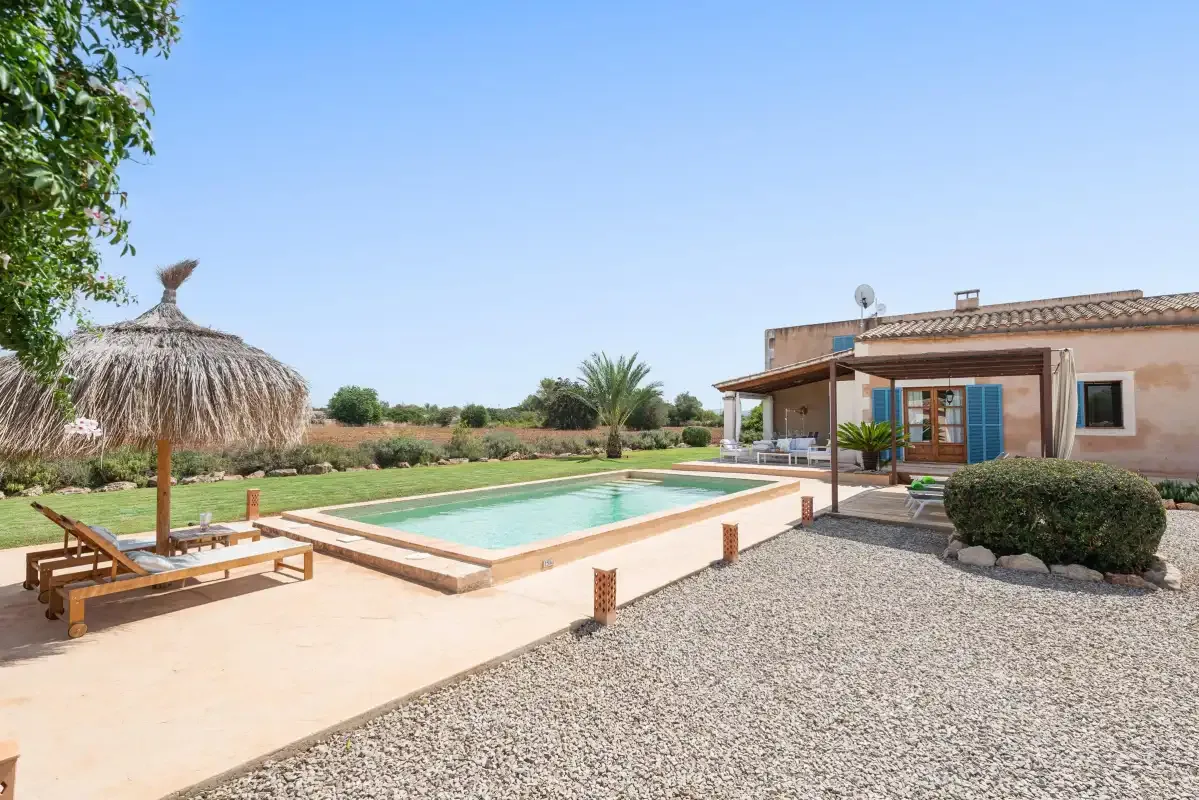 An outdoor swimming pool beside a house, with a thatched parasol and lounge seating.