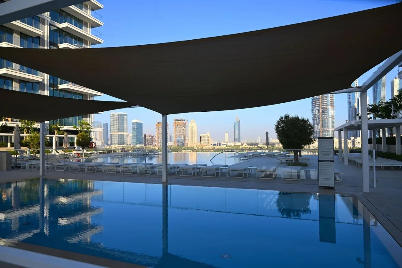 Una piscina al aire libre está bajo amplios toldos, con edificios de la ciudad al fondo.