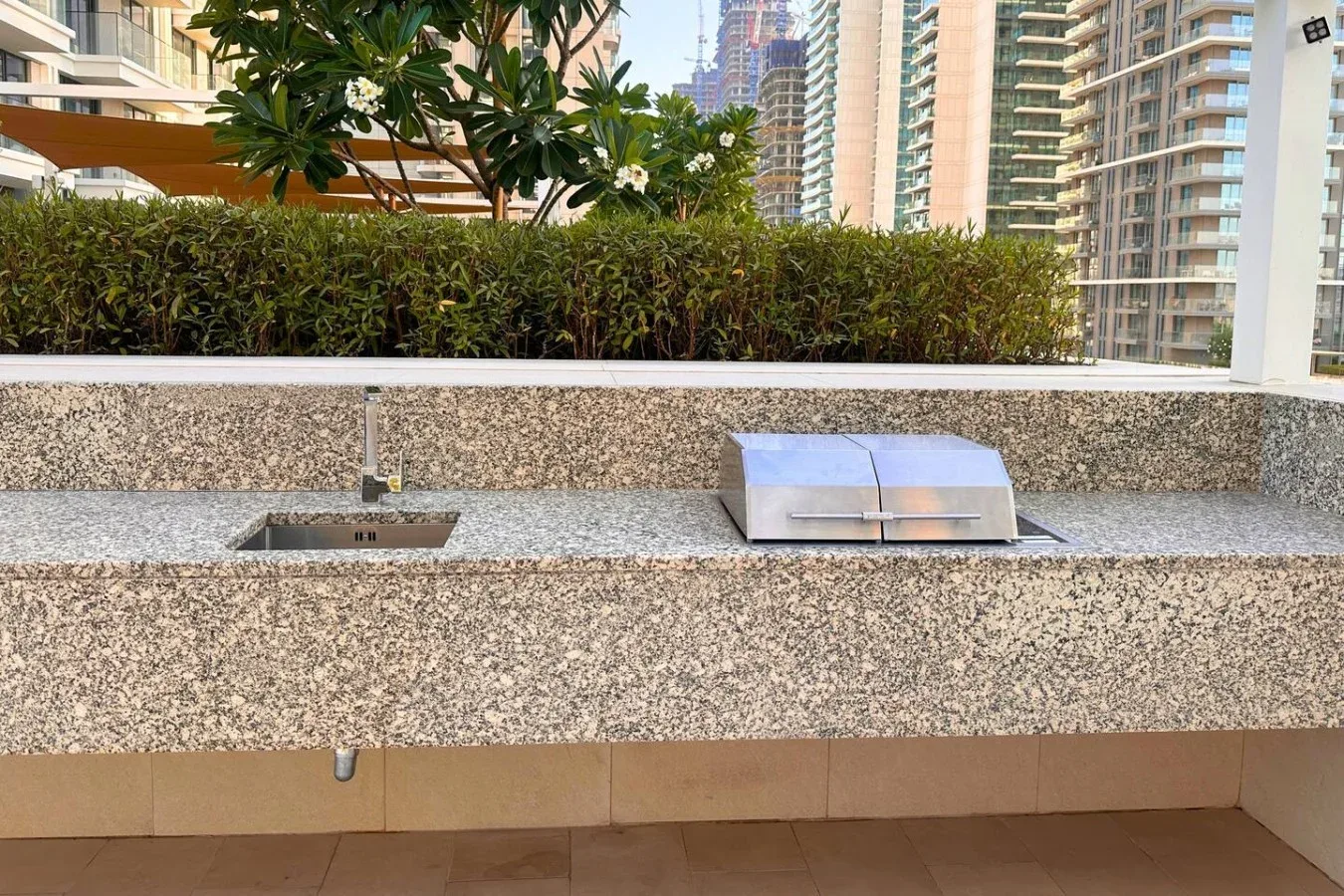 Una encimera exterior de piedra con grifo y lavabo rectangular está junto a un seto plantado.