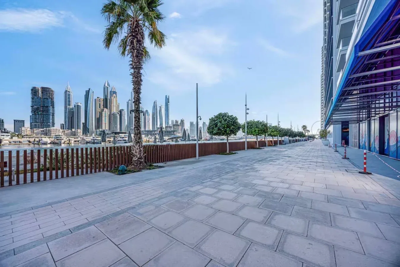 Un paseo pavimentado junto al agua bordea un edificio moderno, con el perfil urbano al fondo.