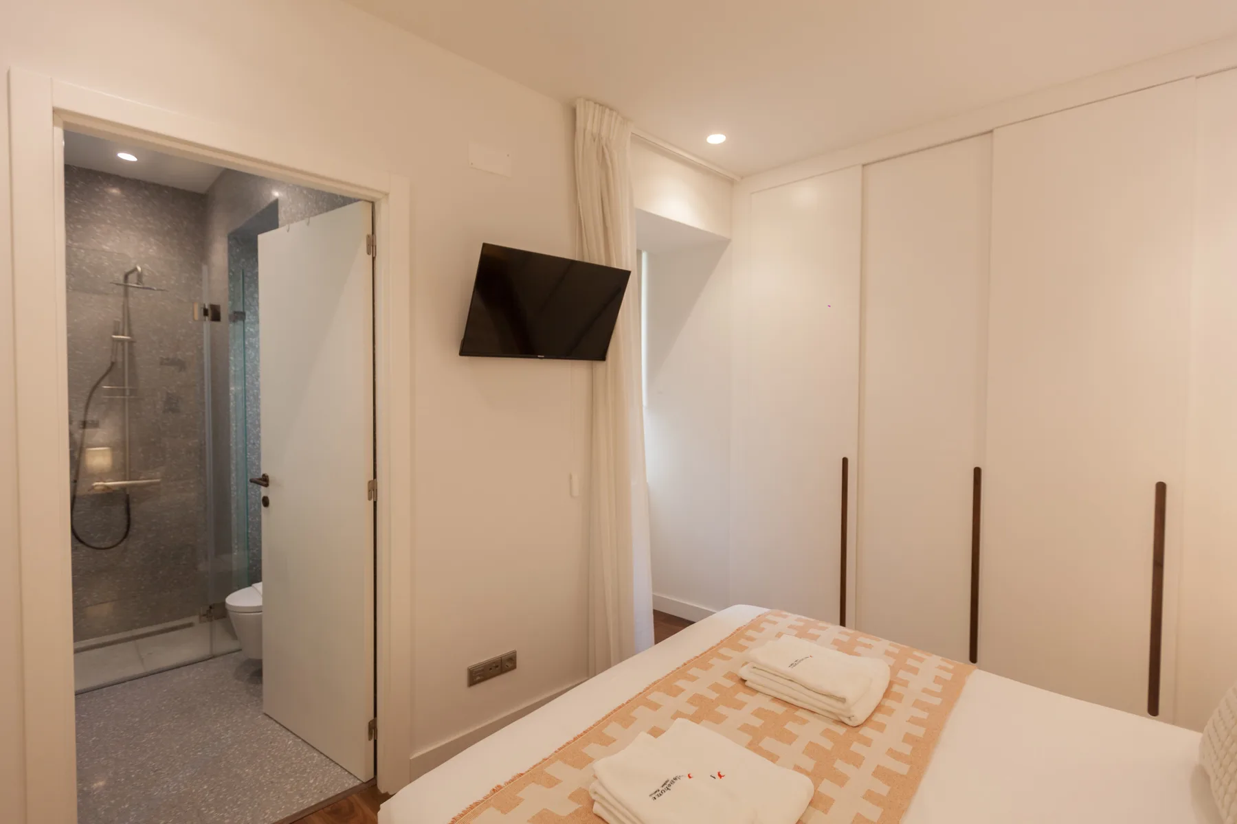 Un dormitorio con una cama, un televisor en la pared y la puerta del baño abierta.