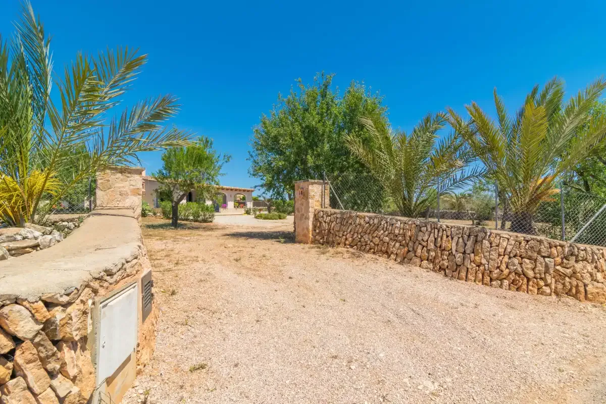 Un camino de grava soleado pasa entre muros de piedra hacia una casa.