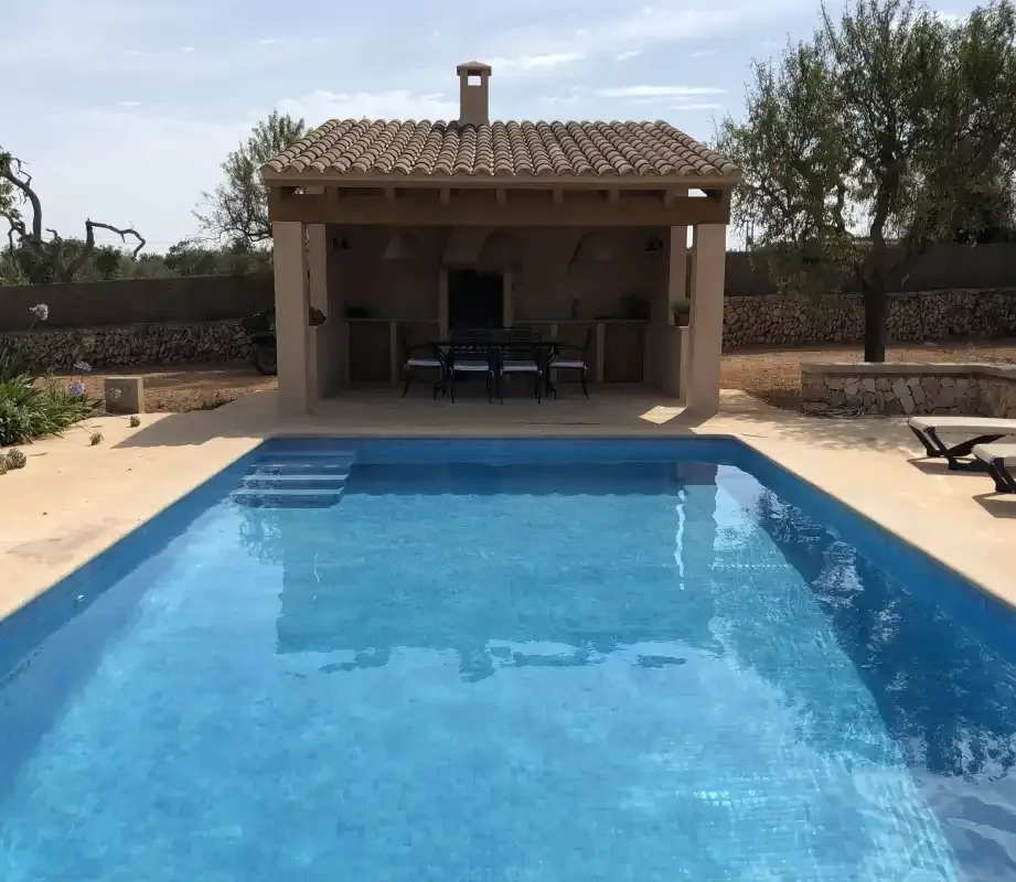 Una piscina al aire libre frente a una zona de comedor cubierta junto a un muro de piedra.