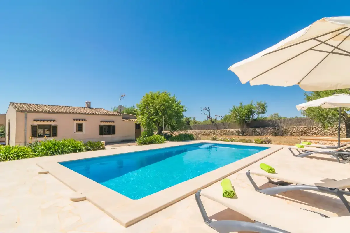 Una piscina exterior soleada junto a una casa baja y asientos de descanso.