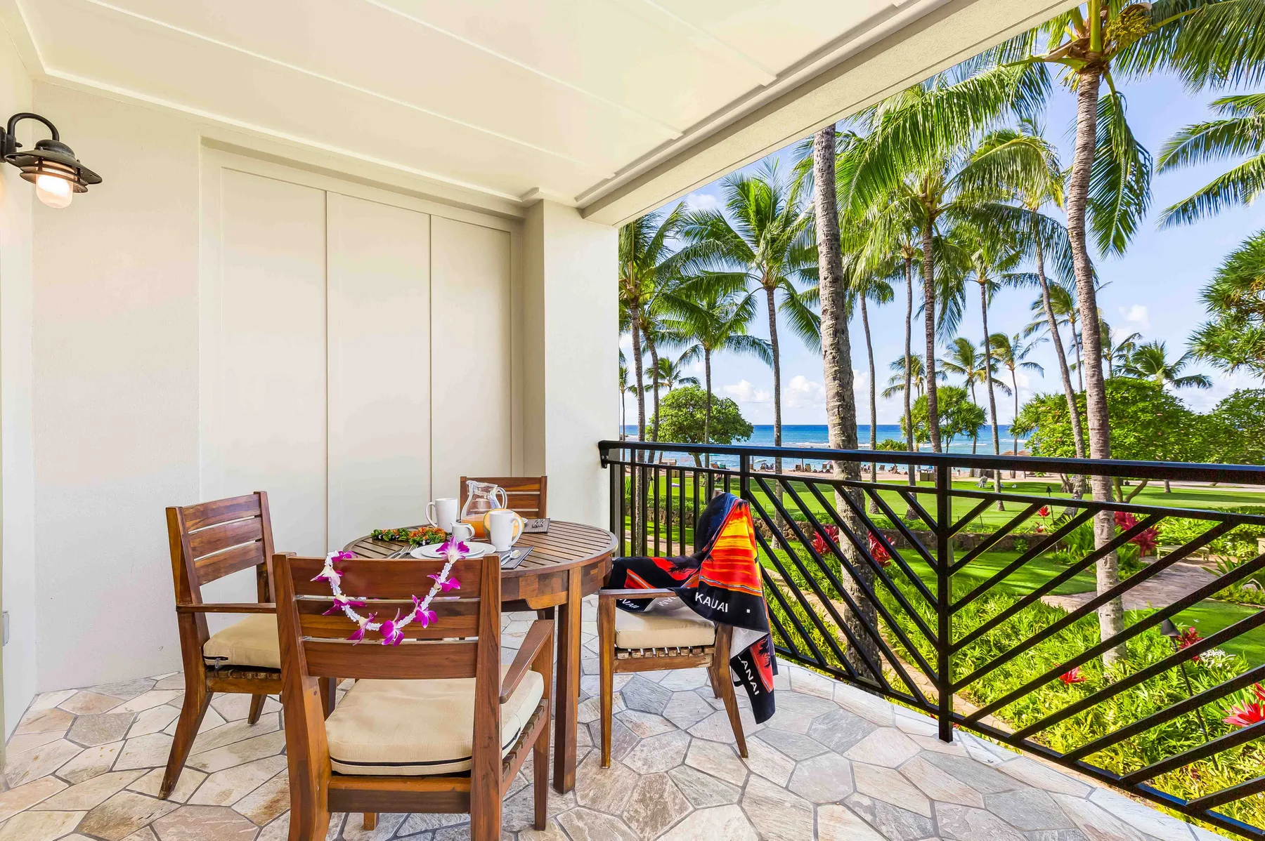 A covered balcony with a dining table overlooks palm trees and the ocean.