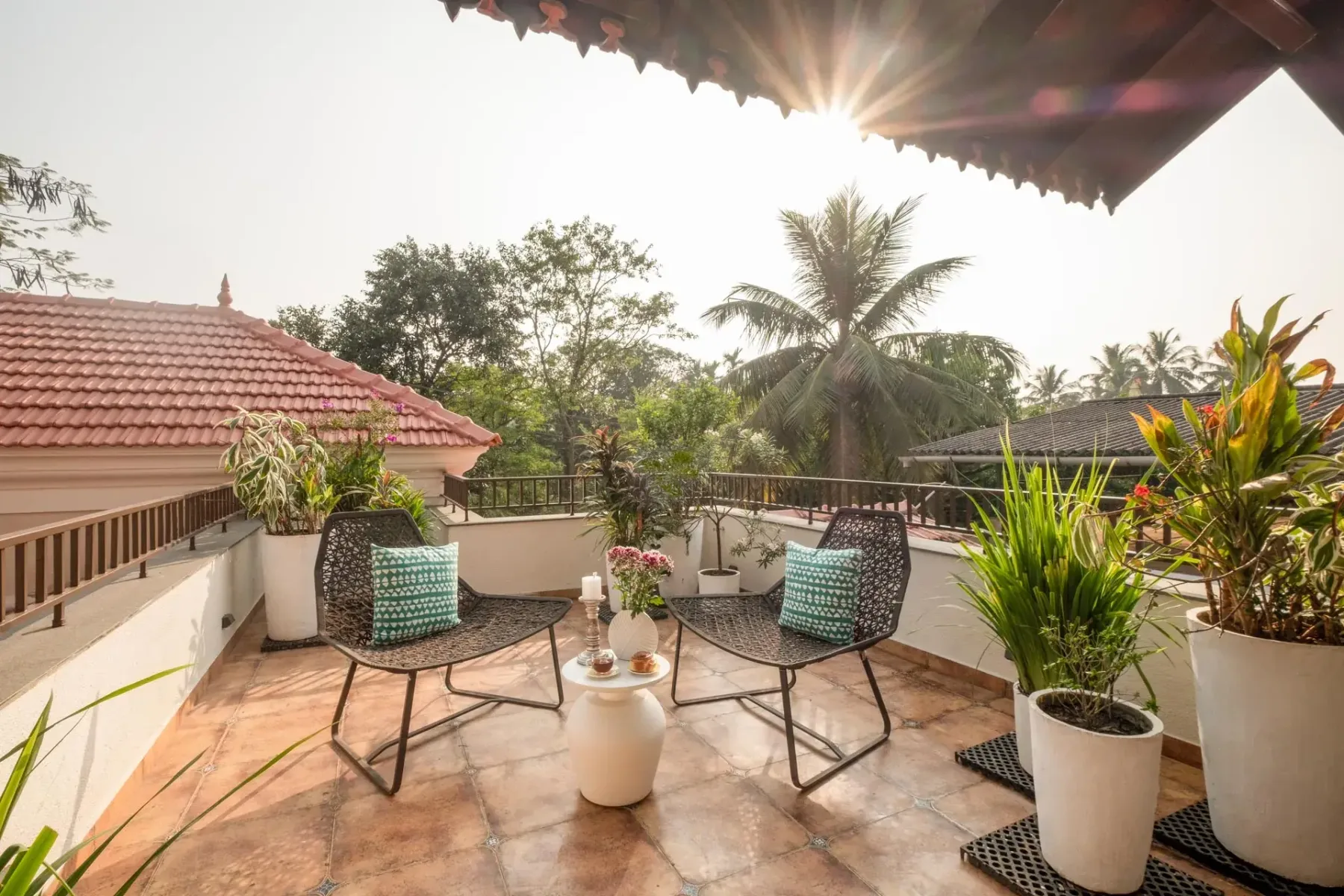 A sunny tiled terrace has two woven chairs, a small table, potted plants, and a view of palm trees.