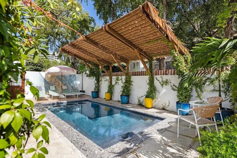 Una piscina al aire libre rodeada de plantas tropicales, con una pérgola de madera que da sombra a las tumbonas.
