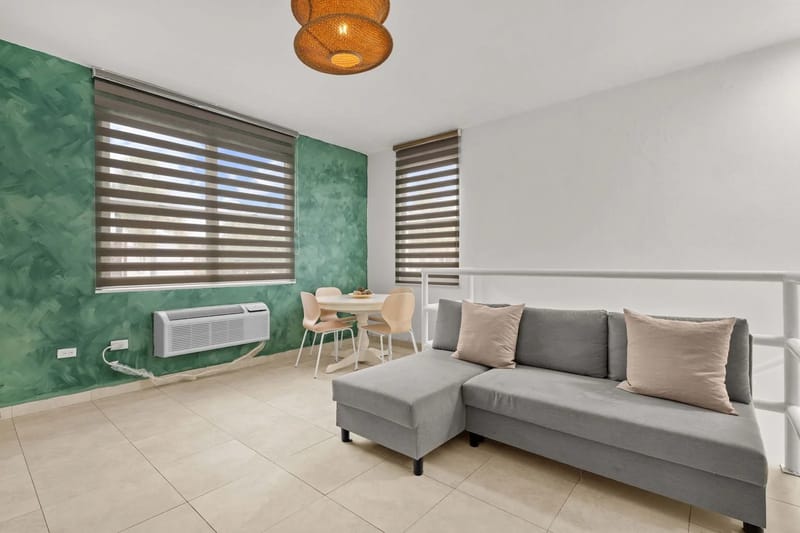 Sala de estar de concepto abierto con sofá seccional, mesa de comedor, ventanas y unidad de aire acondicionado.