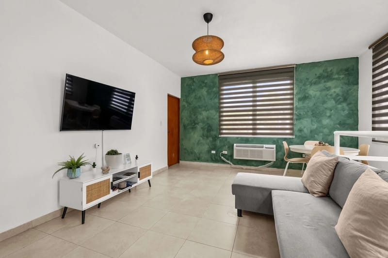 Sala de estar con televisor de pantalla plana montado en la pared, sofá y área de comedor.