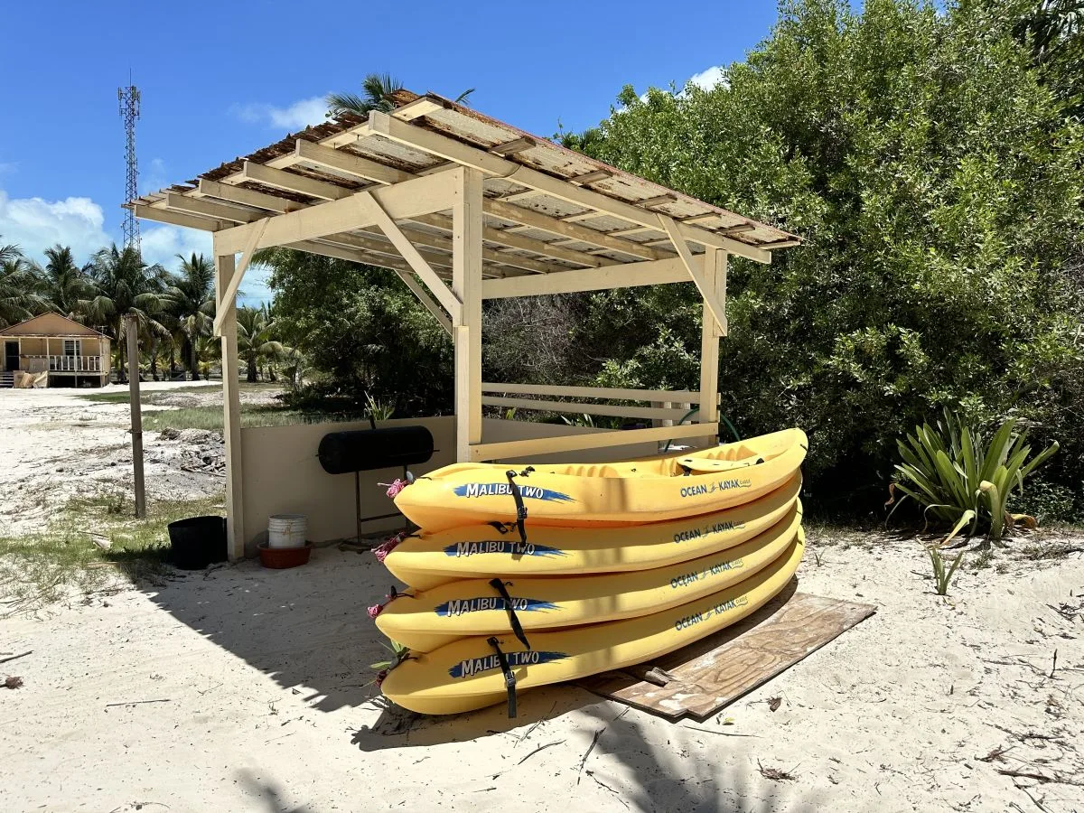 A wooden shelter stands over stacked kayaks on sandy ground.
