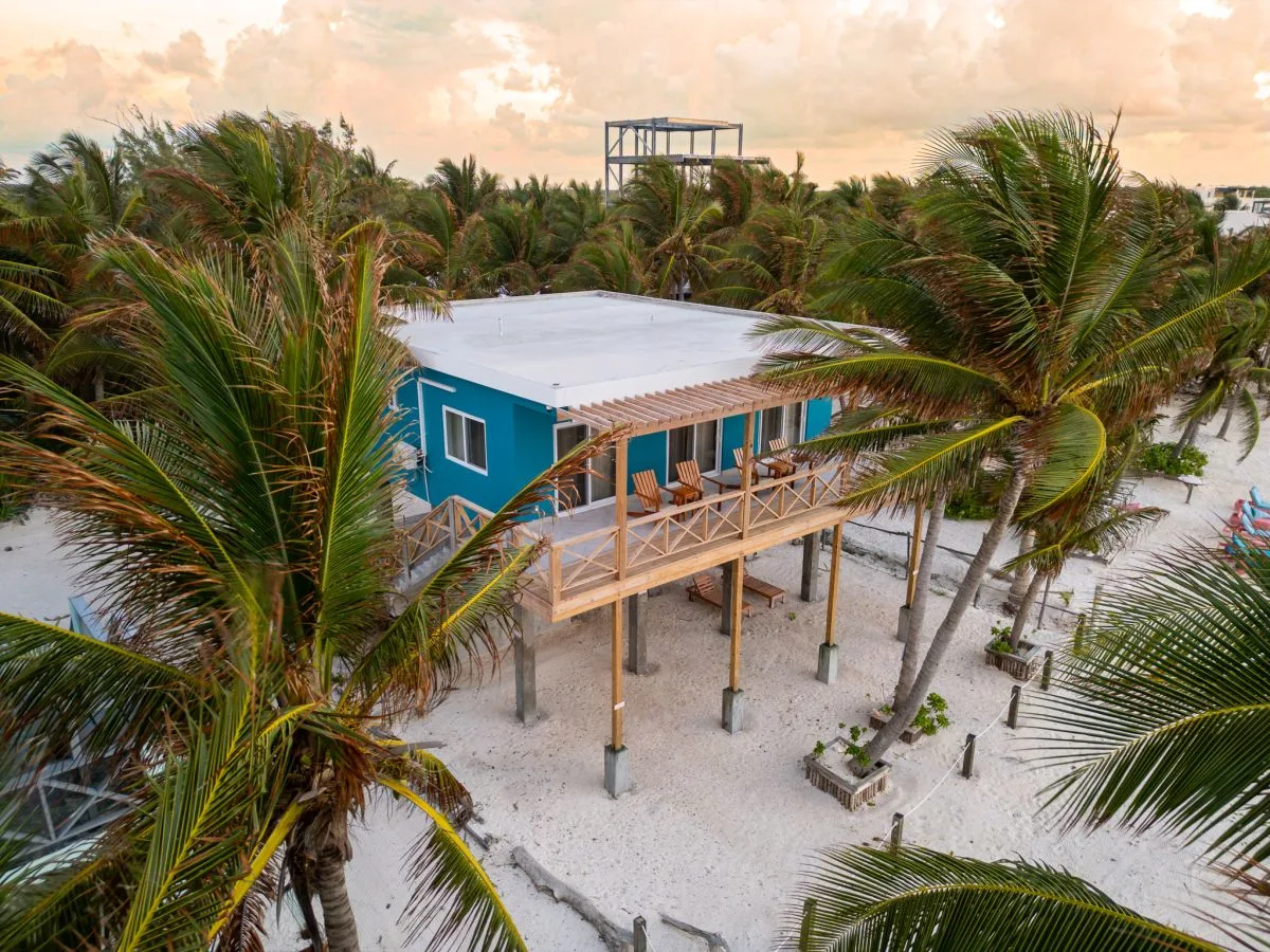 A raised beach house with a long wooden deck stands among palm trees.